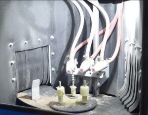 Three nozzles with pink tips and connected tubes are mounted inside a metal machine, aimed at two cylindrical objects on an indexing turntable; the setup appears industrial and automated.