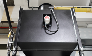 A black metal industrial unit with a red and white electrical socket mounted on top, connected by a black cable. Pipes and other building infrastructure are visible in the background, suggesting this equipment may be part of a Mechanical Recovery System commonly used in Sandblast Booths such as the MARS2000.