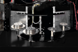 A close-up view of a machine with three metallic rotating platforms, each holding a circular metal object, possibly brake discs; wires and mechanical parts are visible above and around the platforms.