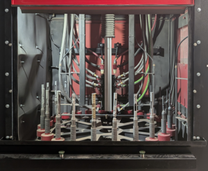 A close-up of industrial electrical or mechanical equipment with multiple wires, metal rods, and cylindrical components inside a metal enclosure. The setup appears complex, with red, black, and gray elements.