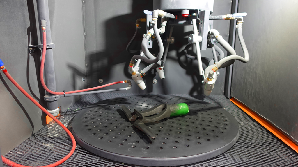 A black metal part with a green end is placed on a circular, perforated platform inside an automation-ready sandblast cabinet, surrounded by robotic arms and tubes.